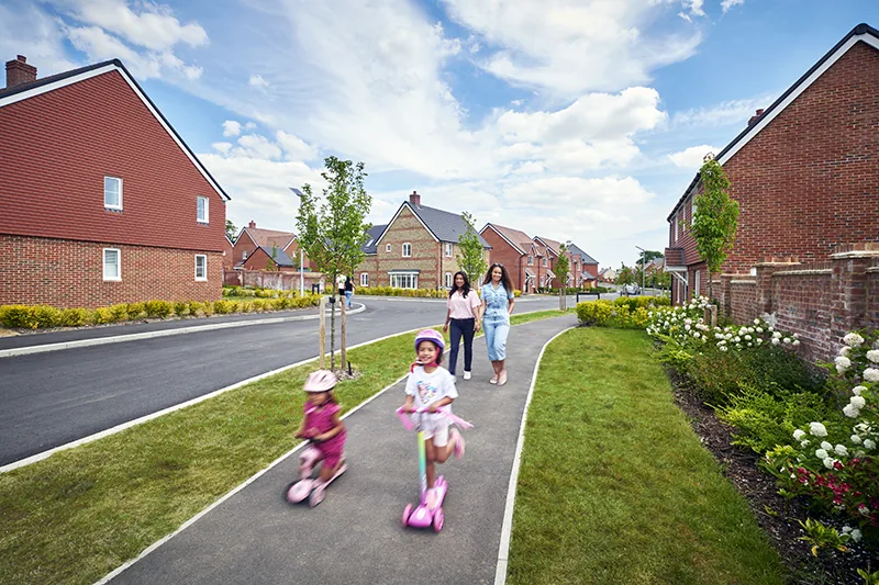 who-we-are Families walking and children riding scooters along a footpath through a new housing development